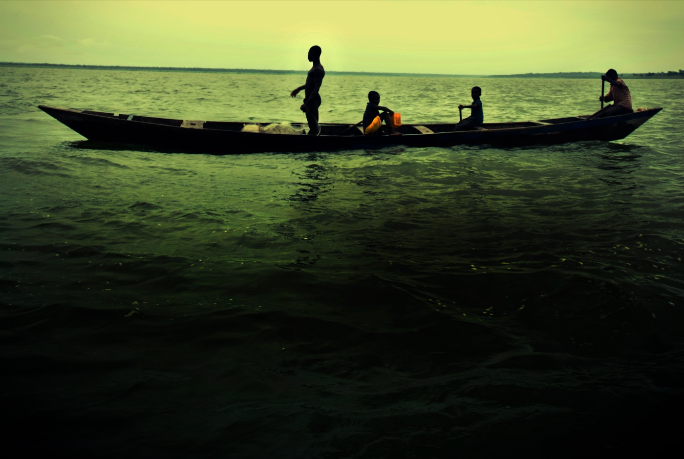 Silhouetted fishermen and children in a long wooden canoe on Lake Volta, Ghana, at dusk.